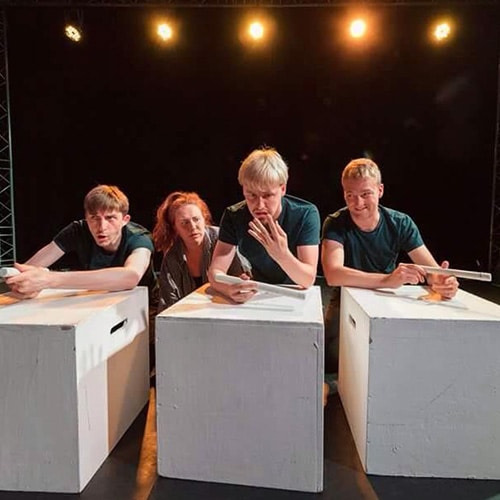 Four actors on stage leaning over white boxes with lights in background